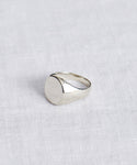 The Gemma Signet Ring, featuring a plain silver band with a round, flat top, is shown on a white textured fabric background.