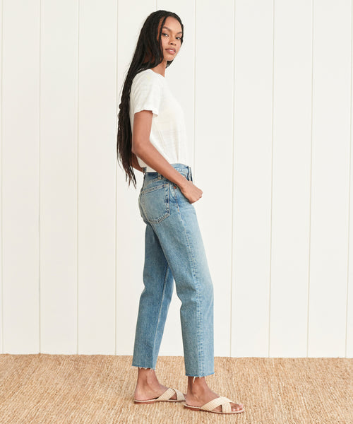 A woman with long braided hair, wearing a white t-shirt, High Rise Stove Pipe Jean, and sandals, stands sideways on a woven rug against a white paneled wall.
