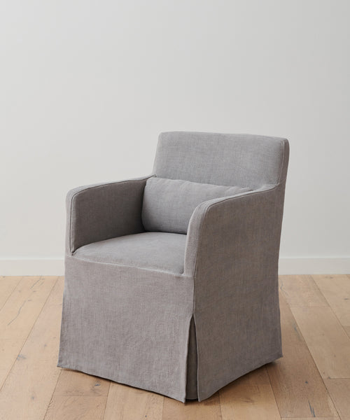 The Sunset Dining Arm Chair Slipcover in light grey ash linen fabric, with a matching lumbar pillow, covers a modern armchair placed on a wooden floor against a plain white wall.