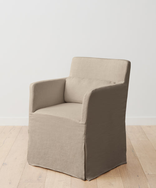 The Sunset Dining Arm Chair, featuring taupe upholstery, a boxy silhouette, straight arms, and a matching lumbar cushion, is placed on a light wooden floor against a plain white wall.