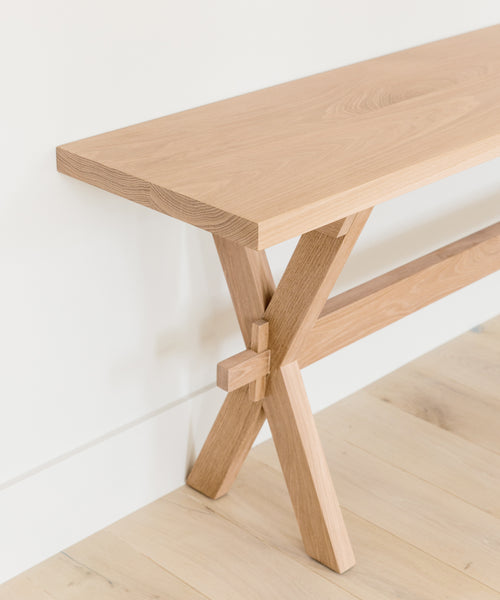 The Oak Entry Table features a minimalist design with a light wood finish and crossed legs, inspired by Jenni Kayne furniture, and looks clean and modern against a white wall on a light wood floor.