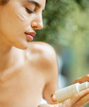 A woman with closed eyes applies cream to her cheek, holding The Brightening Duo bottle. She looks relaxed as softly blurred greenery in the background enhances the serene skincare moment.