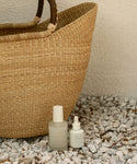 Two Oak Essentials bottles from The Rescue Edit, including Ritual Oil with a dropper, rest on white pebbles beside a woven straw bag—your essentials for a luminous glow.
