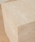 The Sur Side Table, a large beige travertine block with clean edges, sits on a light wooden floor and is illuminated by soft natural light.