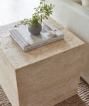 The Sur Side Table displays a gray vase with green foliage and the book Pacific Natural At Home, positioned beside a cream-colored sofa.