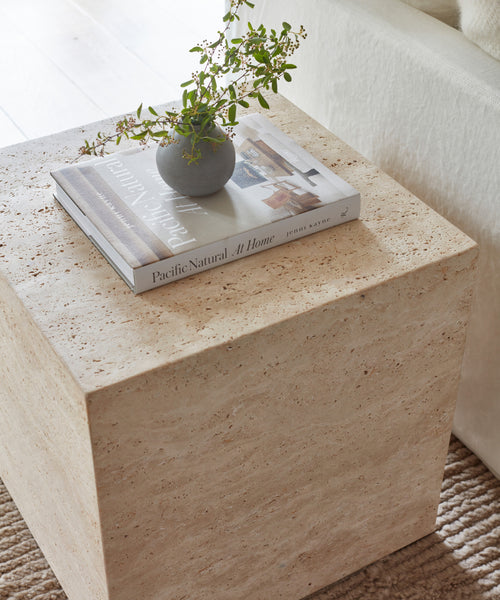 The Sur Side Table displays a gray vase with green foliage and the book Pacific Natural At Home, positioned beside a cream-colored sofa.