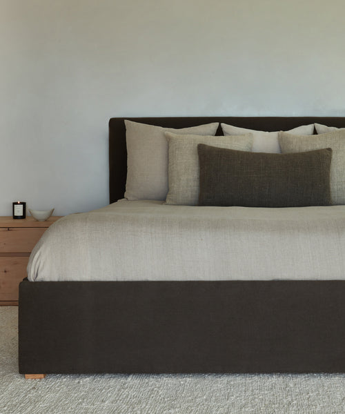 The Pacific Bed features beige and dark brown Belgian Libeco linen bedding, multiple pillows, and a dark brown upholstered headboard beside a wooden nightstand with a candle and bowl, set against a light wall.