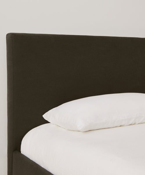 A neatly made Pacific Bed with a dark, upholstered headboard and a single white pillow in luxurious Belgian Libeco linen, set against a plain light-colored wall.