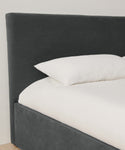 A close-up of a modern bed with a headboard and frame upholstered in Fabric Swatch Slate Linen, featuring a white mattress and pillow, set against a light-colored wall.