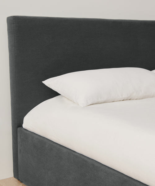A close-up of a modern bed with a headboard and frame upholstered in Fabric Swatch Slate Linen, featuring a white mattress and pillow, set against a light-colored wall.