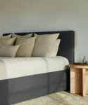 A modern bedroom featuring the Pacific Bed in dark gray Slate Linen with beige and cream pillows, a light bedspread, a wooden side table with vase, and a textured beige rug. Fabric Swatch Slate Linen available upon request.