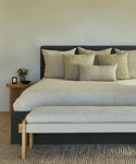 A modern bedroom with a neatly made Pacific Bed featuring a grey upholstered headboard, beige throw pillows, a light bench at the foot, a wooden nightstand with a small plant and candle, and a textured rug on the floor.