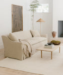 A light, minimalist living room features a beige Miramar Sofa Slipcover, fringed throw, pale rug, low wooden coffee table, ceramic décor, potted plant, rustic lamp, and white walls with a high window.