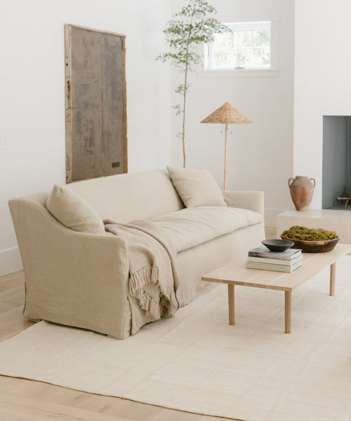 A light, minimalist living room features a beige Miramar Sofa Slipcover, fringed throw, pale rug, low wooden coffee table, ceramic décor, potted plant, rustic lamp, and white walls with a high window.