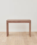 The Newport Desk, featuring two understated drawers, stands against a plain white wall on a light wood floor, bringing a simple yet timeless vintage-inspired look to your space.