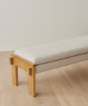 The Ynez Bench, featuring a minimalist white oak design and light cushion seat, sits on a light wood floor against a plain, light-colored wall.